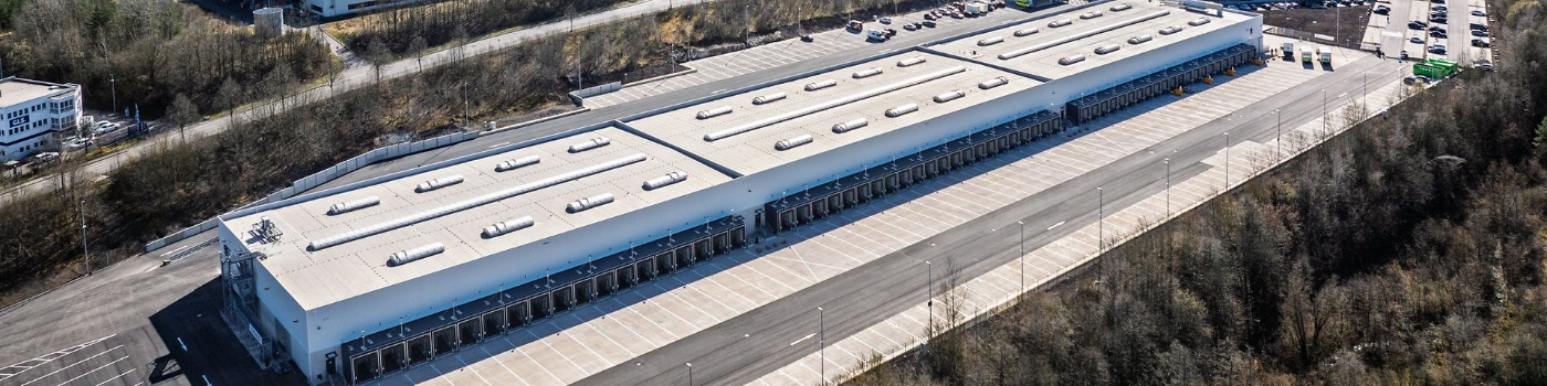 Aerial view of a large DB Schenker logistics warehouse with multiple loading docks and adjacent parking areas, surrounded by trees.