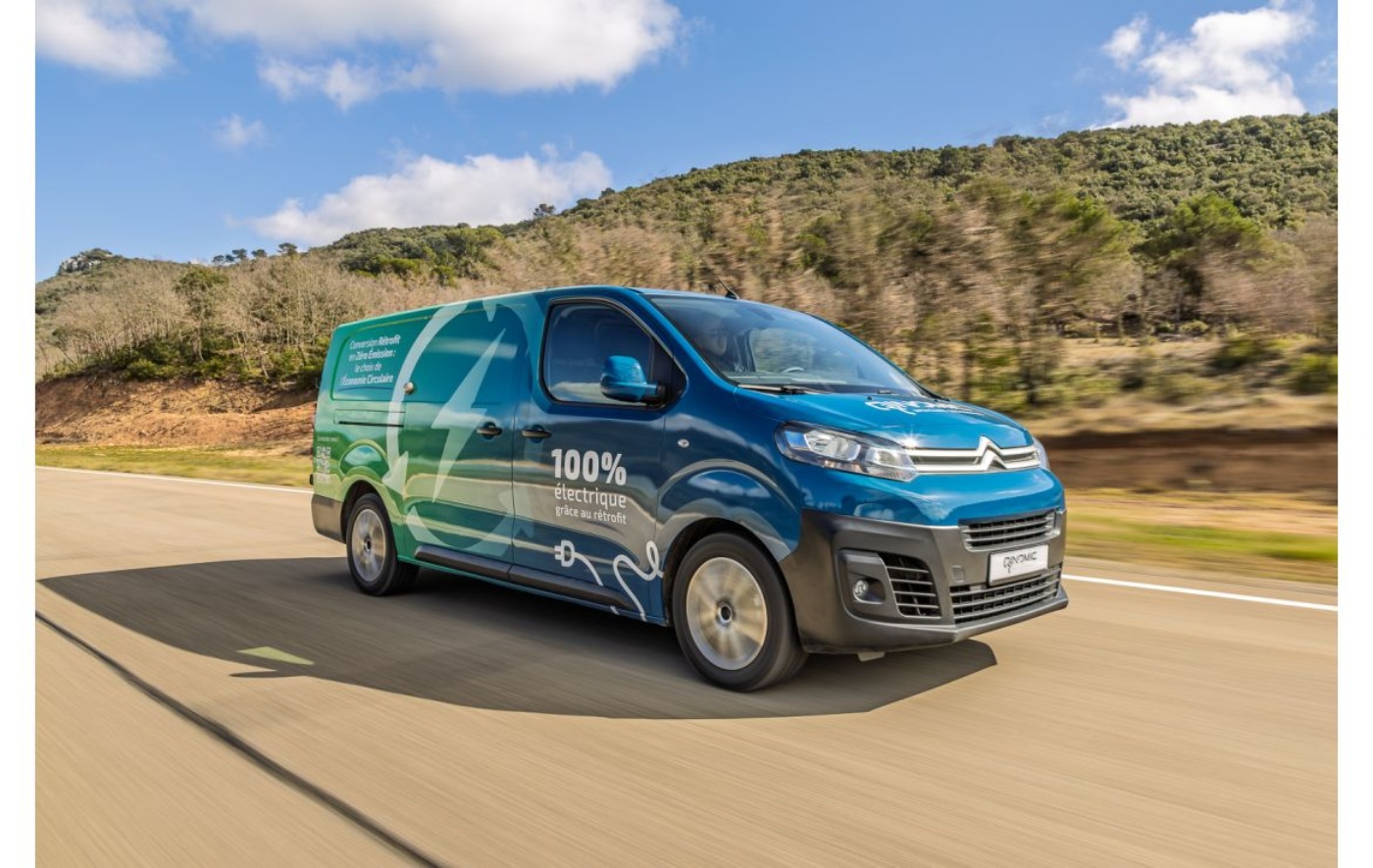 Blue Citroën electric van with "100% électrique" written on the side, driving on a road through a landscape with trees and hills.