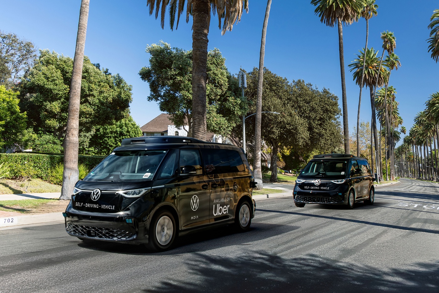 Two black self-driving Volkswagen vans branded with Uber logos driving on a sunny palm-lined street.
