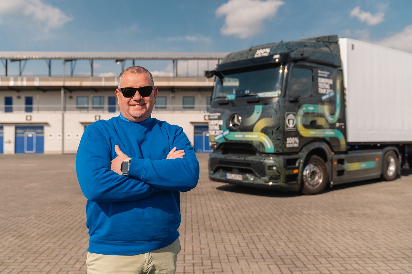Man in a blue sweater and sunglasses standing with crossed arms in front of a modern Mercedes-Benz electric truck in an open industrial area.