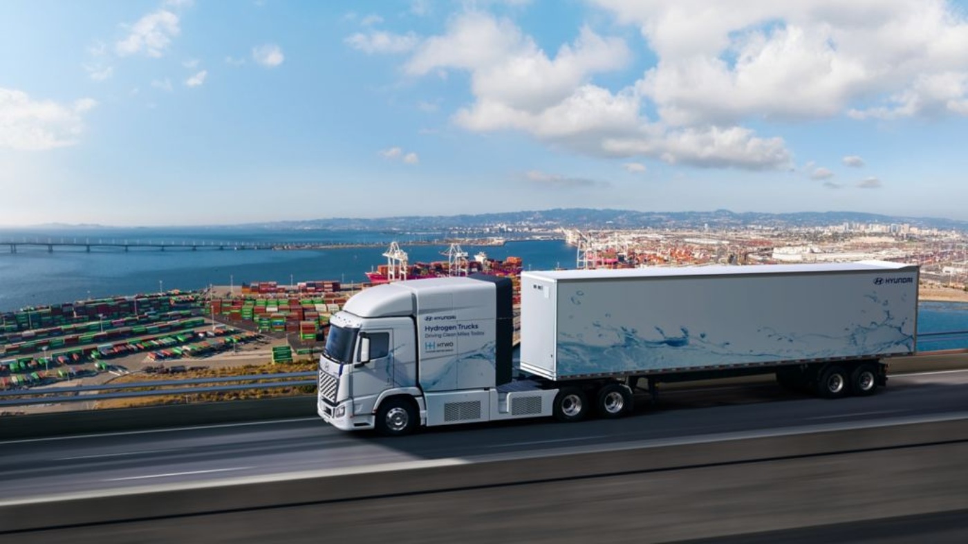 White Hyundai hydrogen truck driving on a road overlooking a busy port area with colorful shipping containers and a bridge in the background.