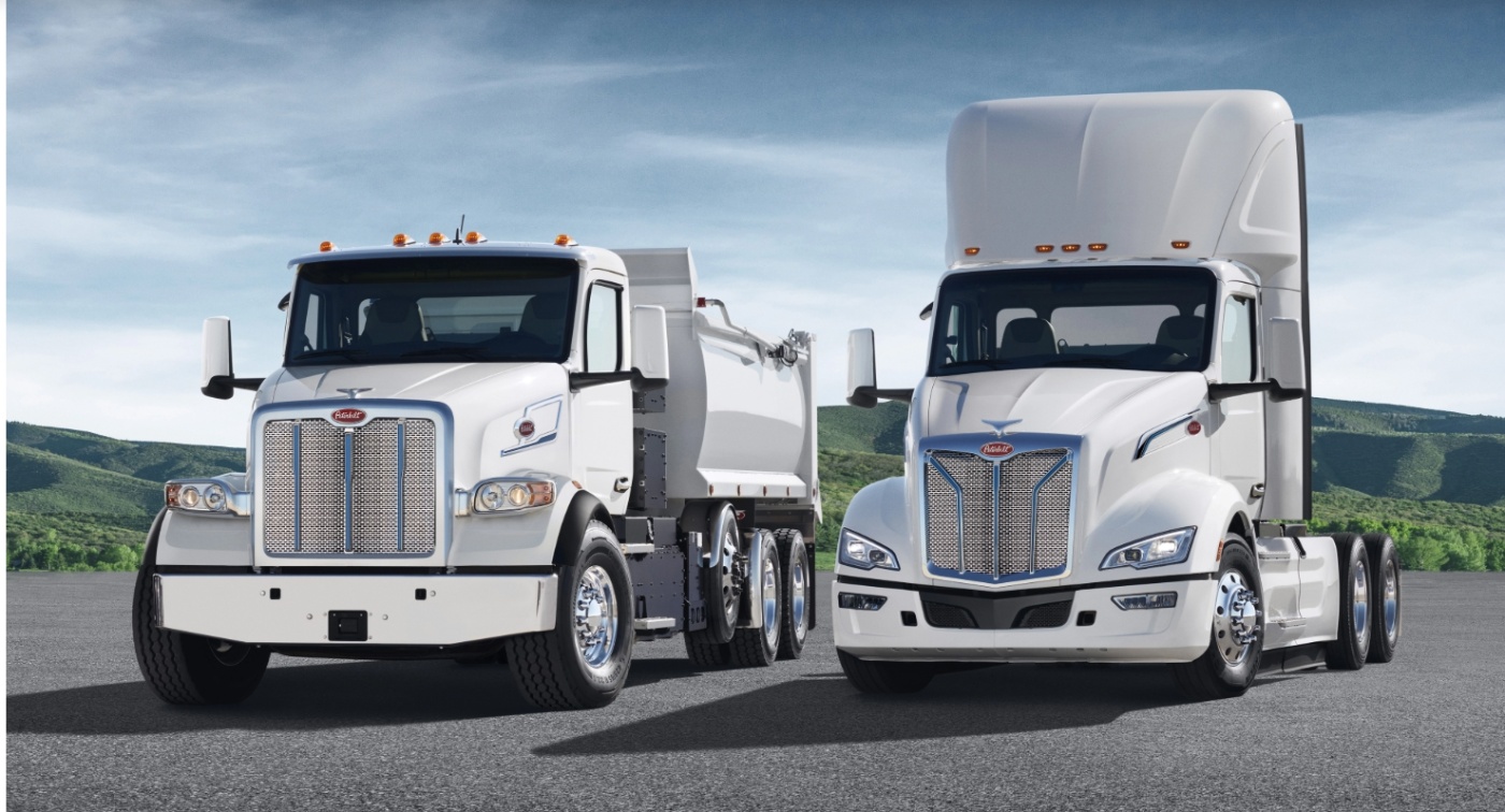 Two white Peterbilt trucks parked side by side on an open asphalt surface, with green hills and a blue sky in the background.