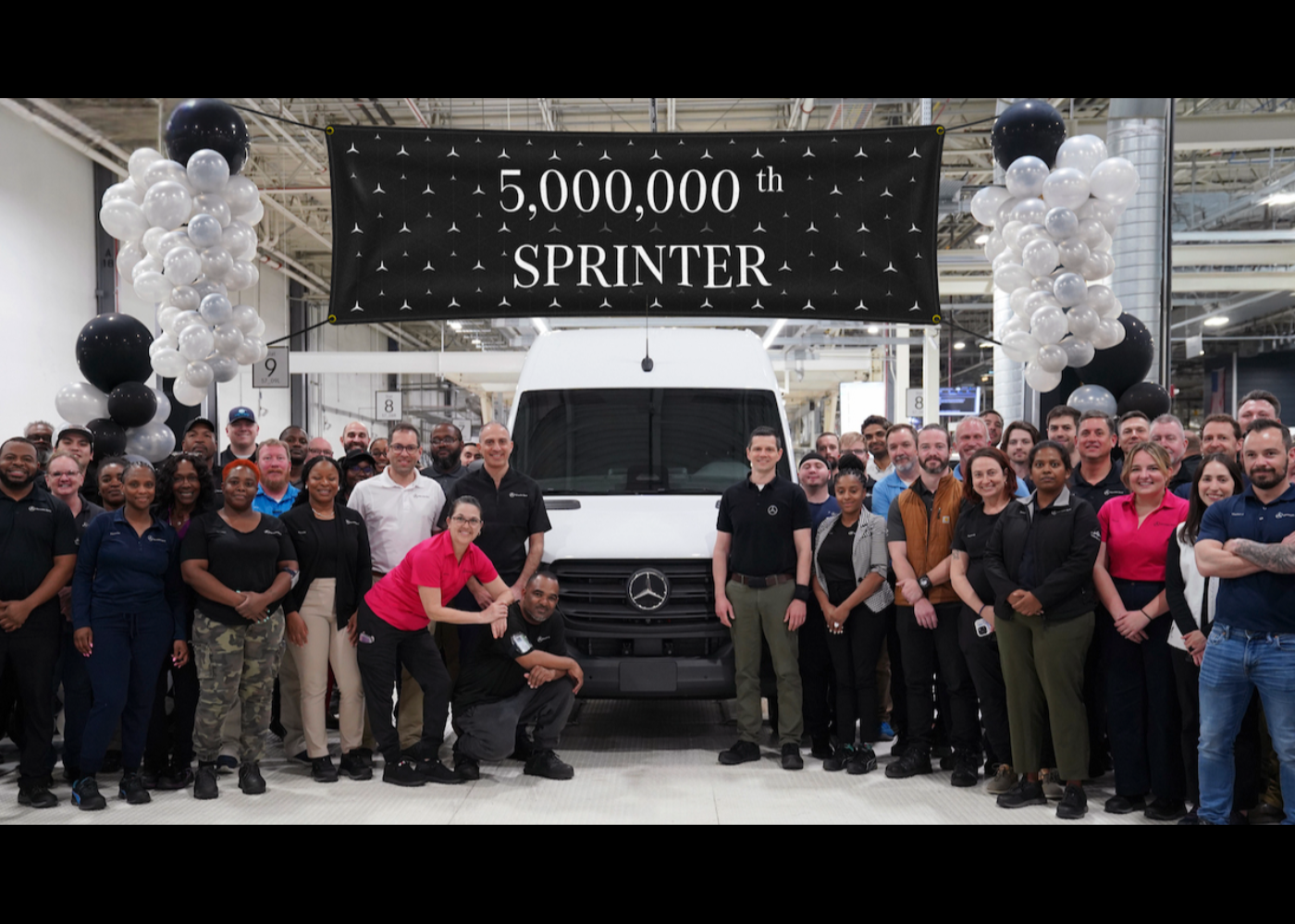 Gruppe von Fabrikmitarbeitern steht vor einem weißen Mercedes-Benz Transporter und feiert die Produktion des fünfmillionsten Sprinters, mit einem schwarzen Banner und Ballondekoration darüber.