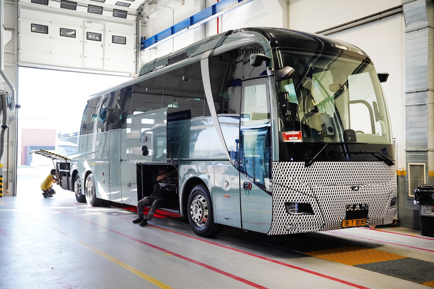 A modern intercity coach is being serviced in a workshop, with two mechanics working in its open storage compartments.