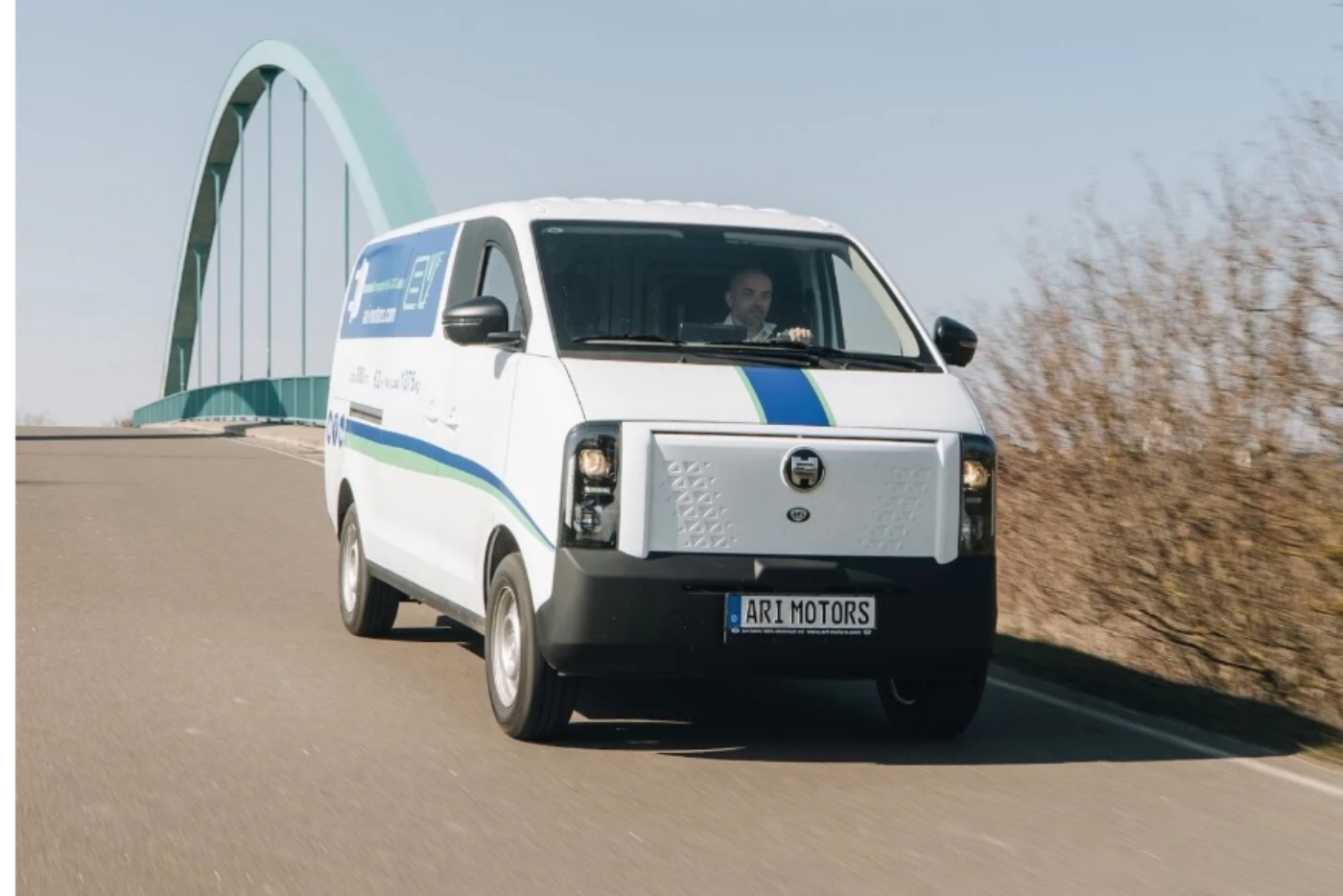 A white electric delivery van from ARI Motors is driving on a road near a bridge, with a driver visible through the windshield.