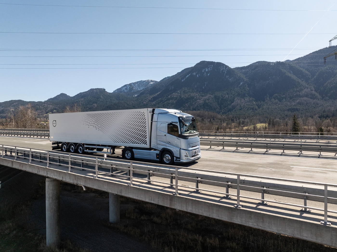 A silver Volvo truck with a modern trailer is driving on a highway bridge, with forested mountains in the background.