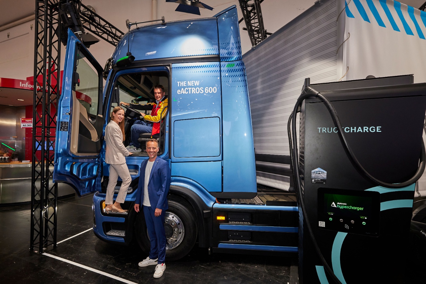 Three people pose with a blue eActros 600 electric truck and a nearby truck charging station.