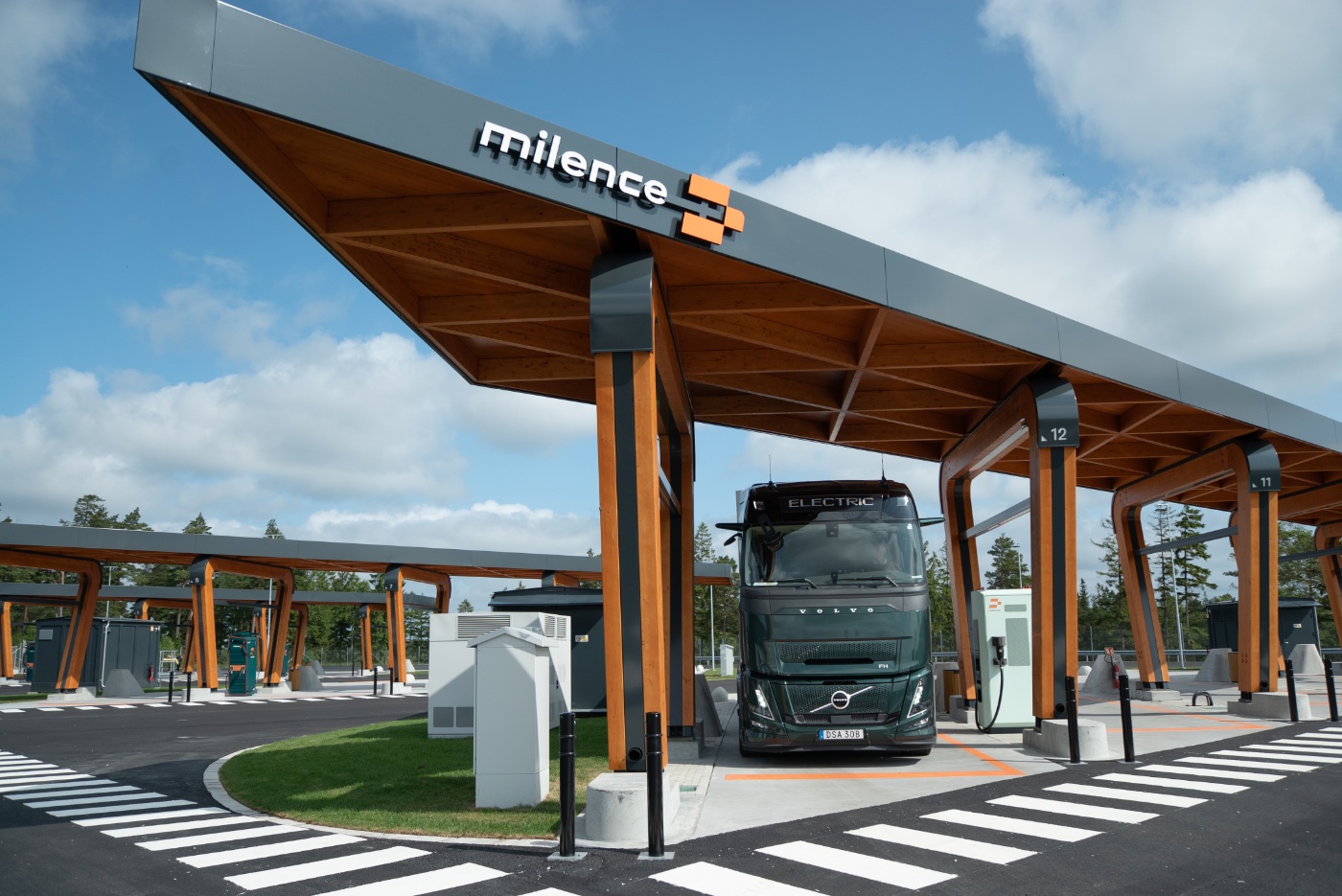 Elektro-Lkw an Milence-Ladestation unter Holzüberdachung bei klarem Himmel.