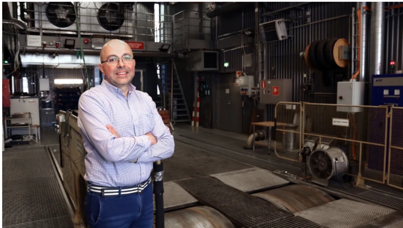 Lächelnder Mann mit Brille steht in einer technischen Prüfanlage mit Geräten im Hintergrund.