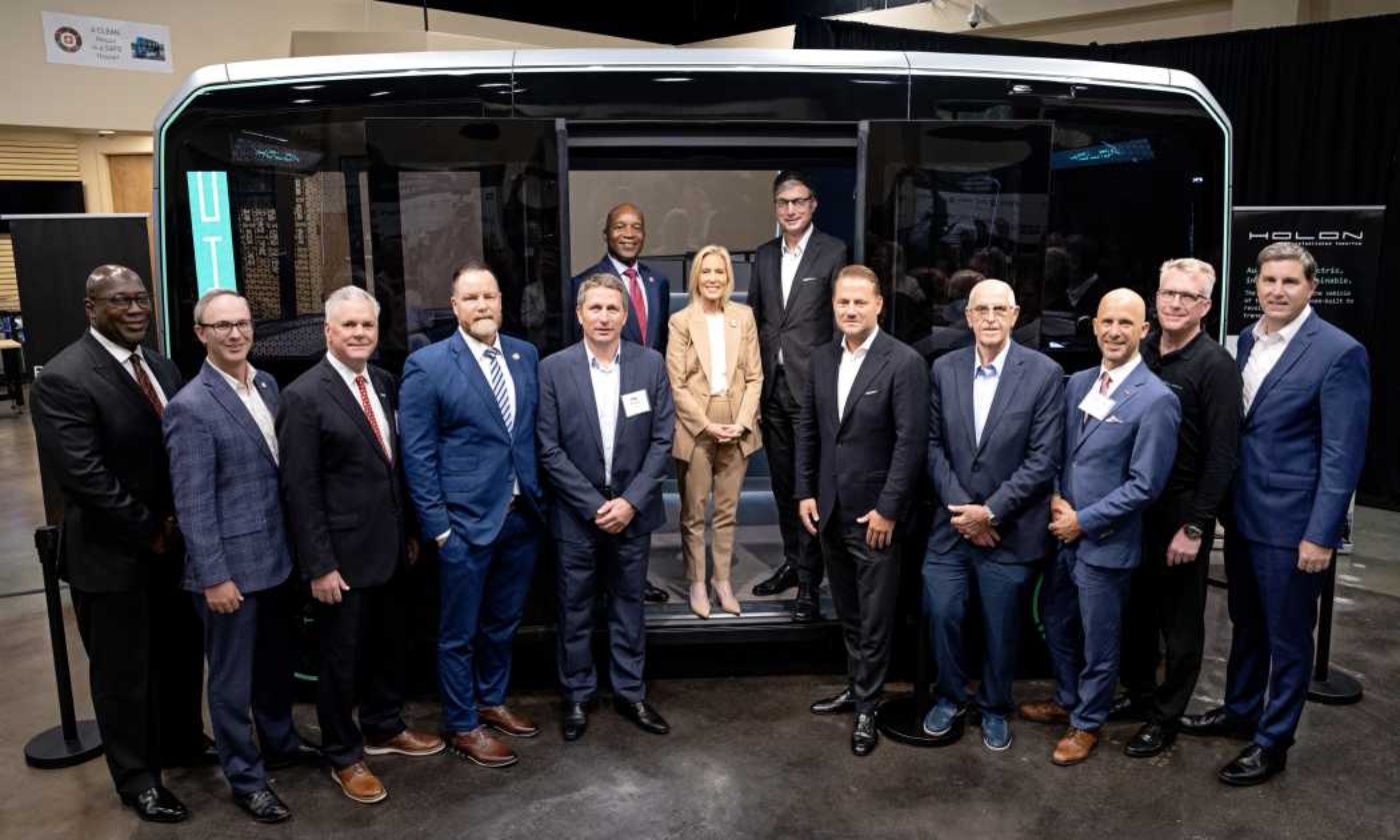 Group of business people standing in front of a modern autonomous shuttle at an indoor event.
