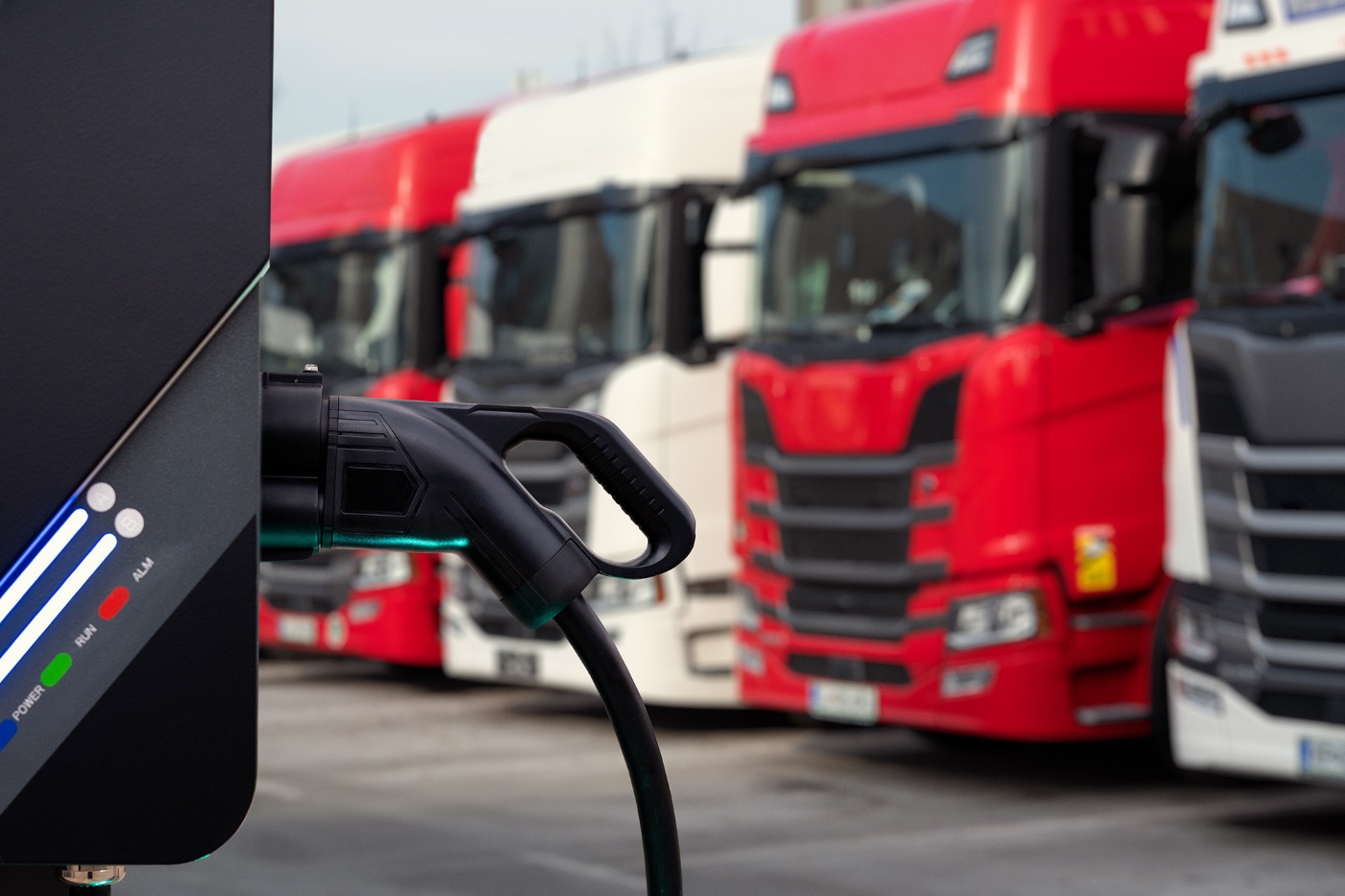 Electric charging plug in front of parked red and white trucks at a charging station.