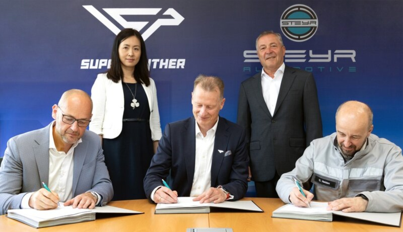 Three men sign documents at a table, while two others stand behind them in front of company logos.