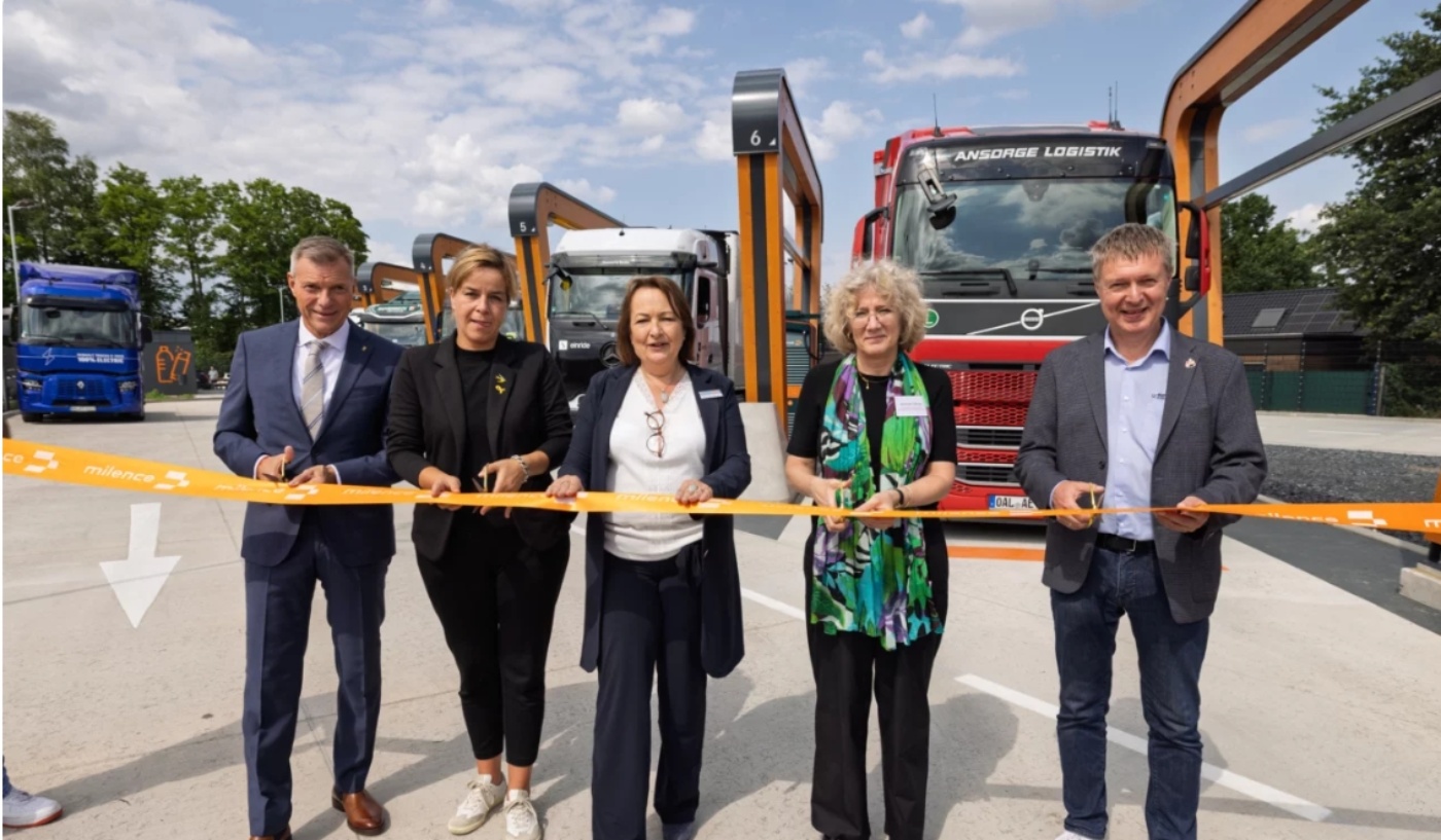 Five people cut a ribbon in front of electric trucks at the opening of a charging station.