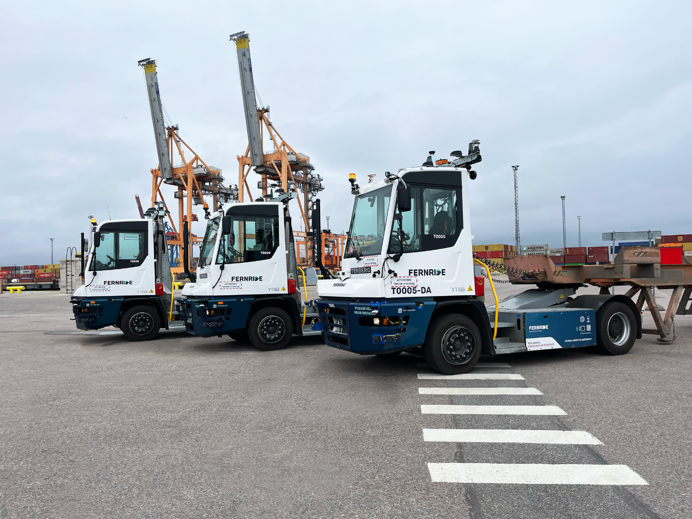Three autonomous yard trucks are parked in a port area with cranes and containers in the background.