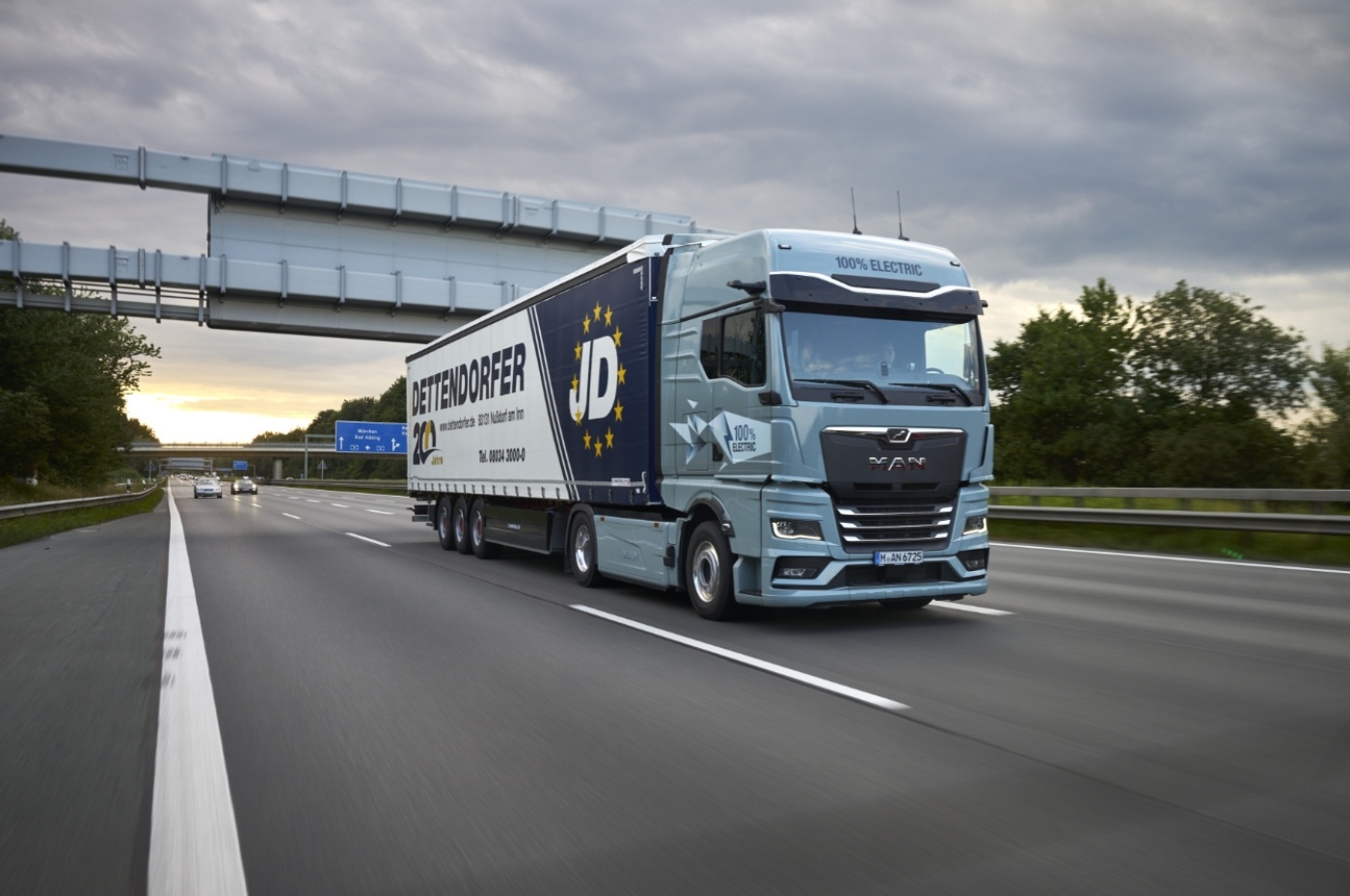 Electric MAN truck with Dettendorfer trailer drives on a German highway under cloudy sky.