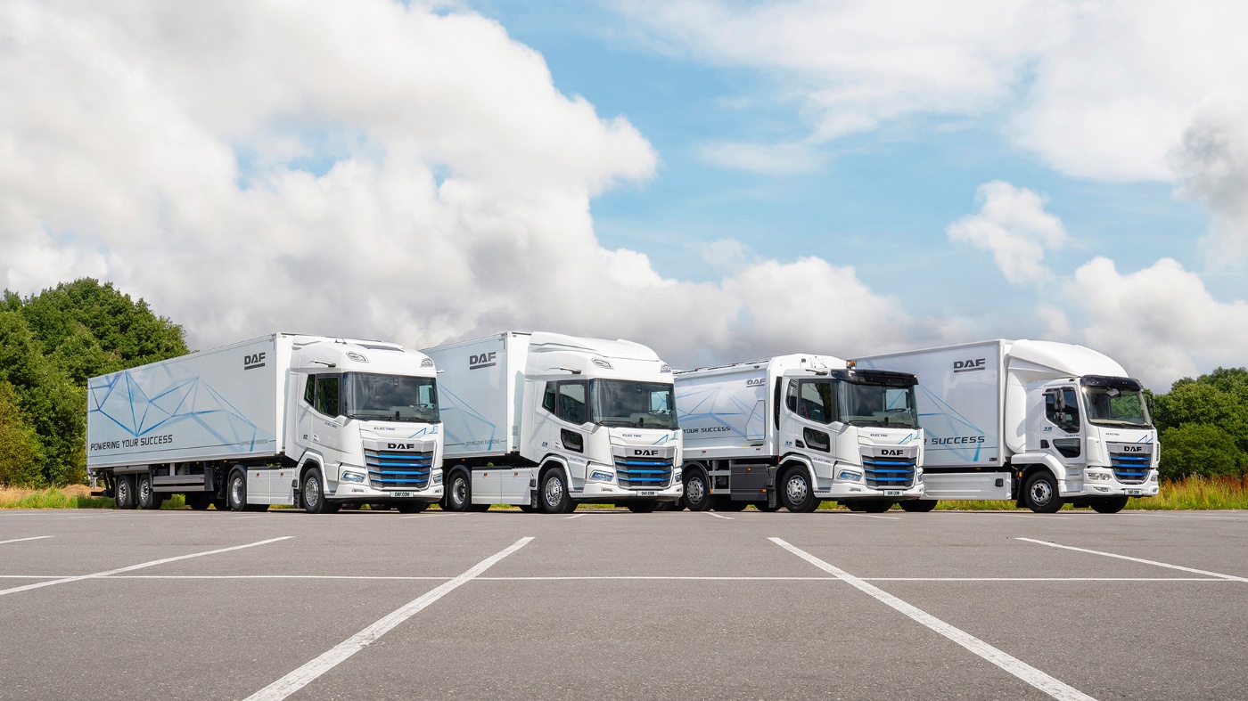 Four white electric DAF trucks are parked in a row on an open lot under a cloudy sky.