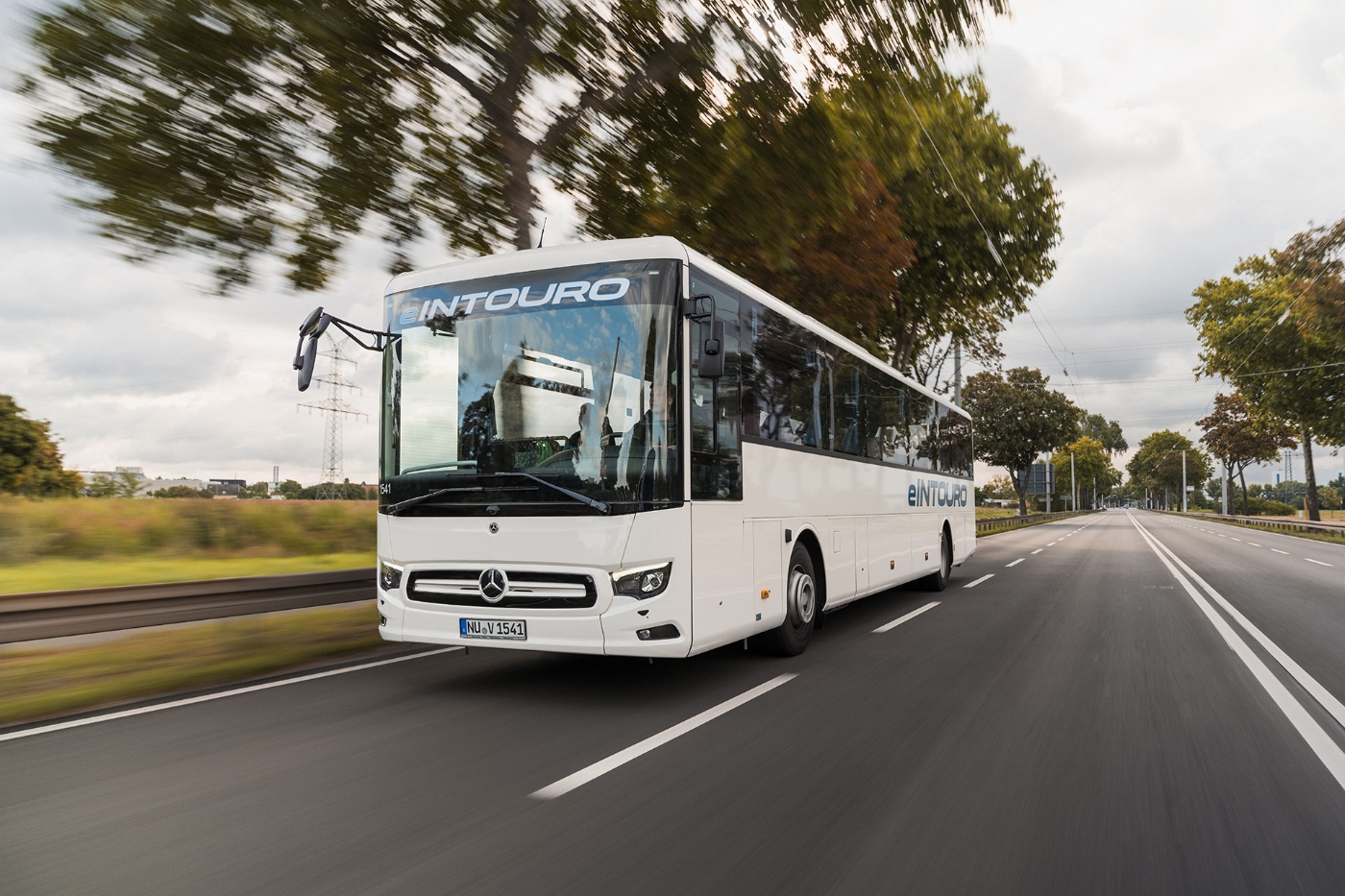 Electric intercity bus with Mercedes logo drives on a tree-lined road.