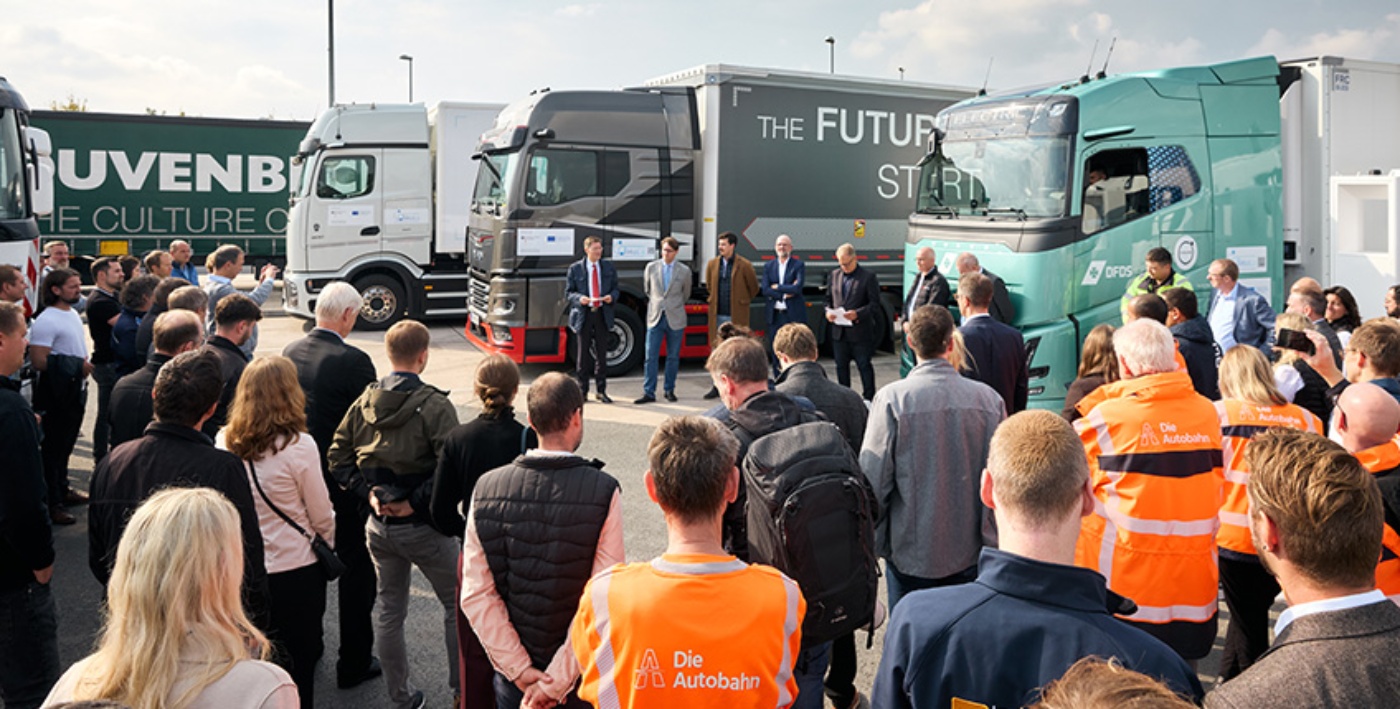 A group of people at a truck event listening to a speaker in front of several branded trucks.