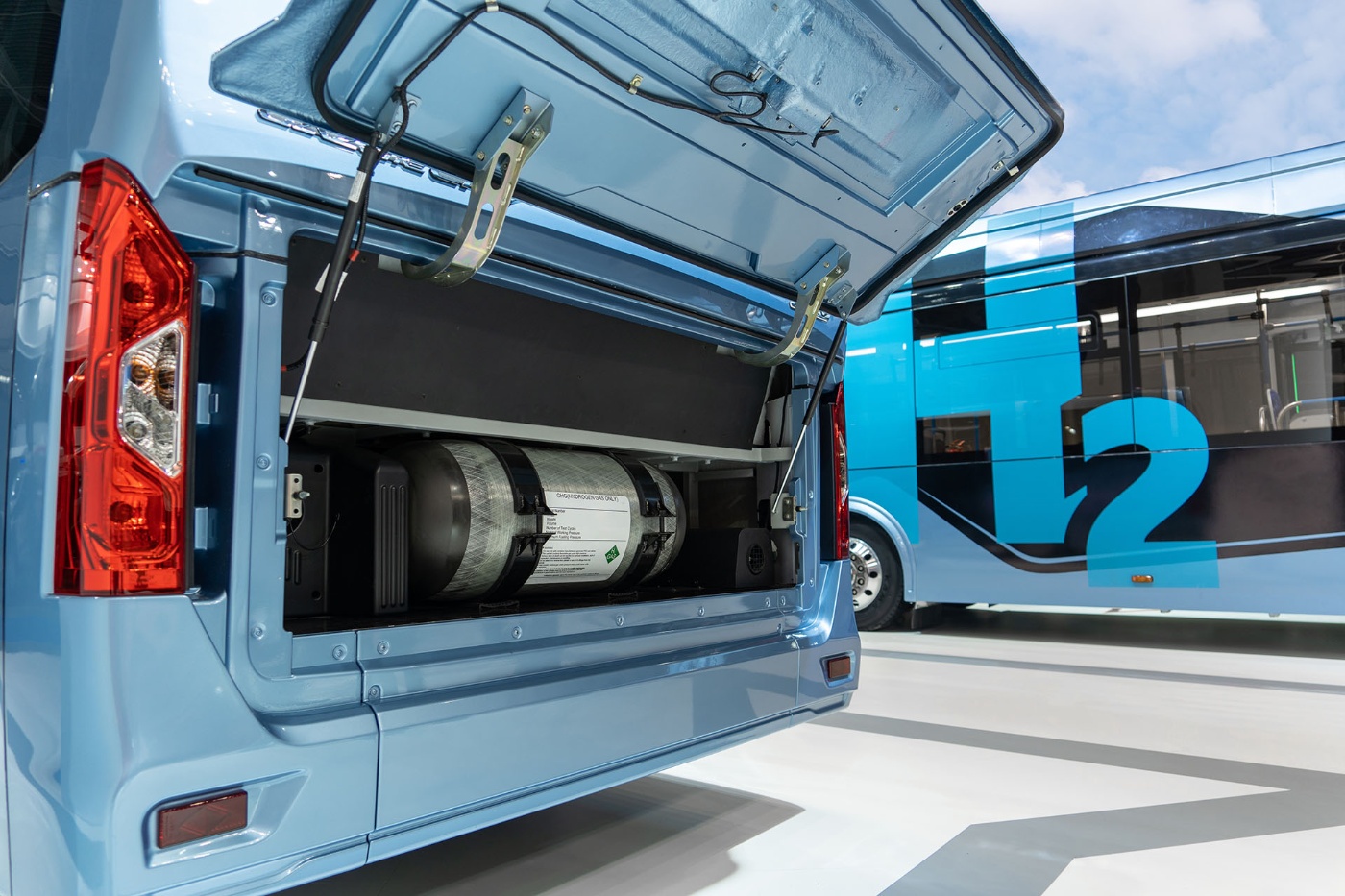 Open rear compartment of a hydrogen vehicle showing the H2 tank, with another bus in the background.