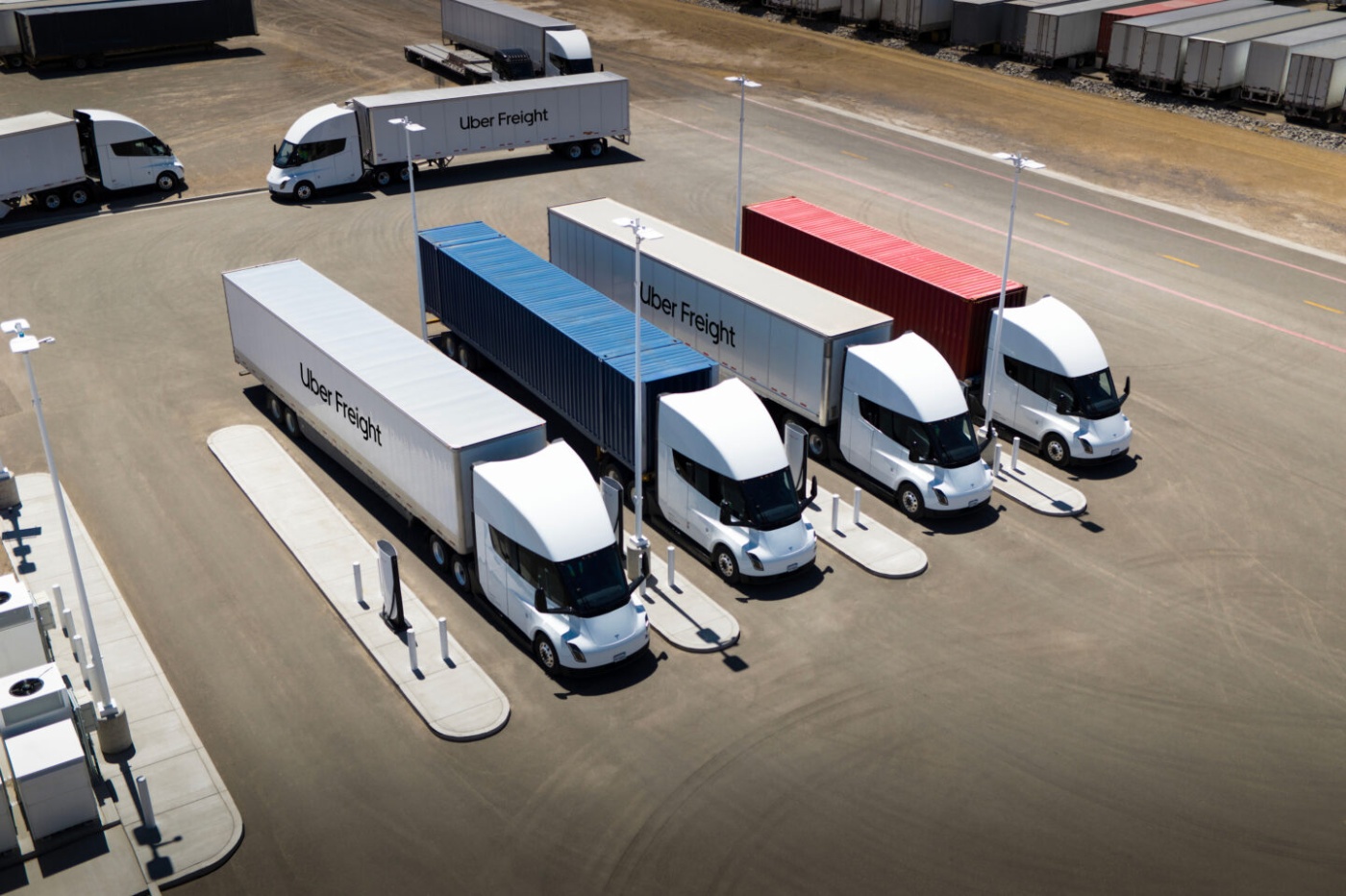 Several electric semi-trucks with Uber Freight trailers are charging at a station in a parking lot.