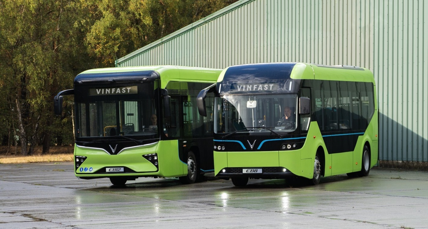Two green VinFast electric buses are parked side by side in front of a green industrial building.