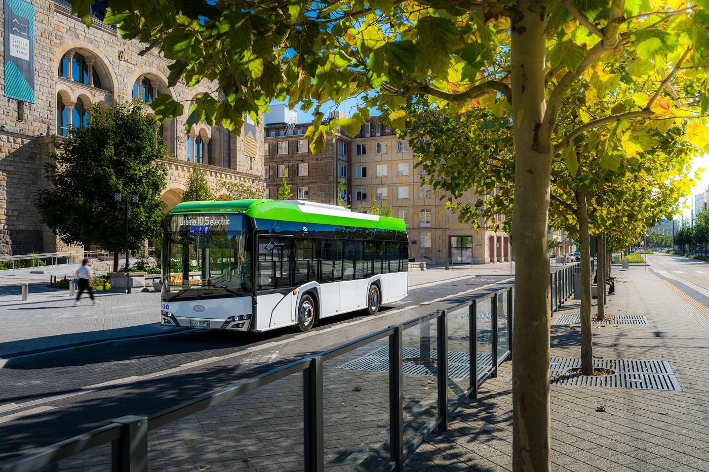 An electric Solaris bus drives through a sunny city street lined with trees and historic buildings.
