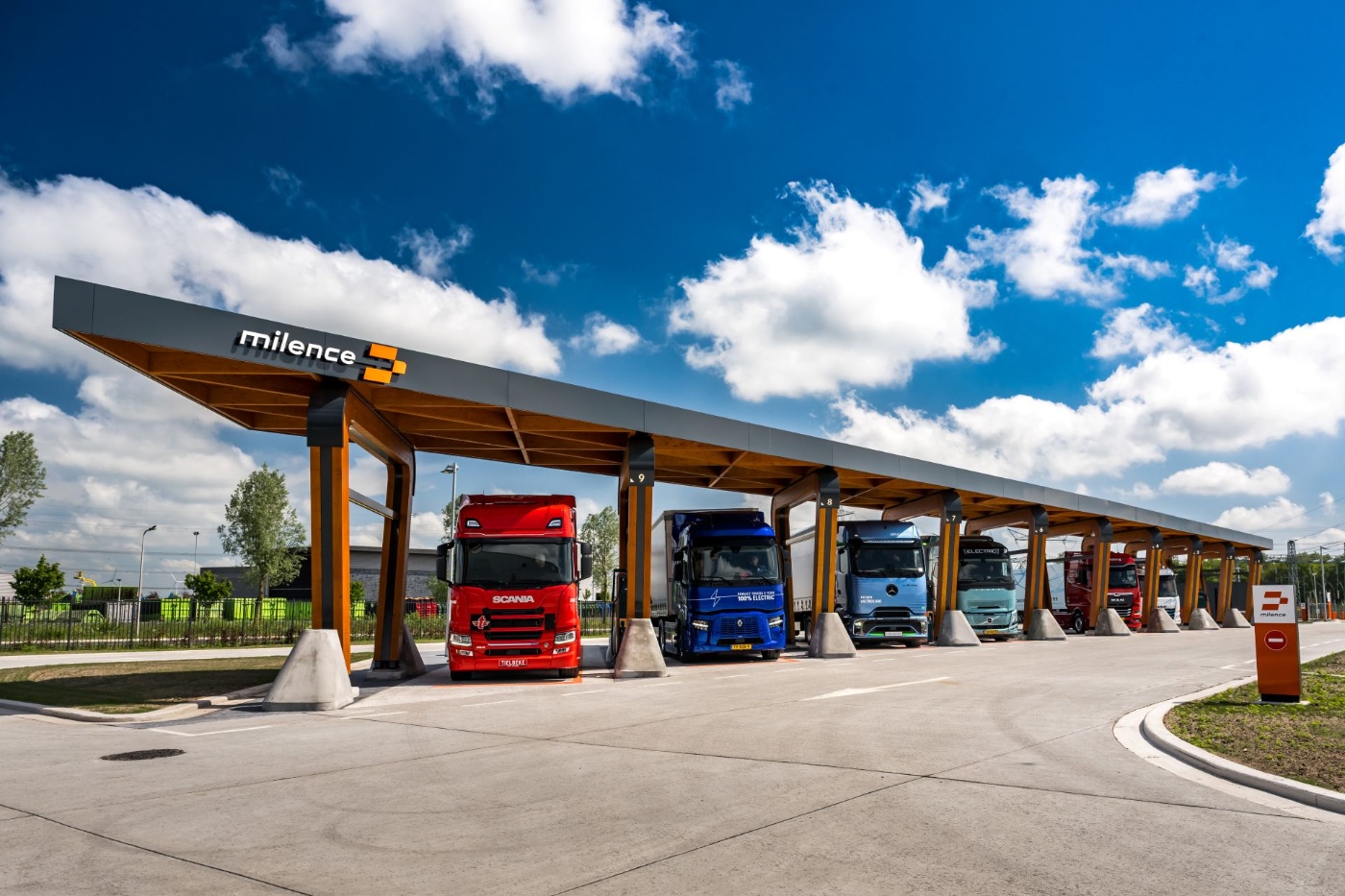Several electric trucks are parked under a covered Milence charging station.