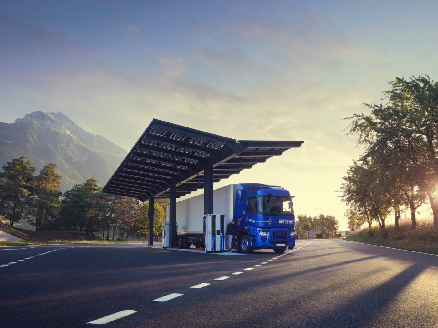 An electric truck charges at a solar-powered station on a scenic road at sunset.