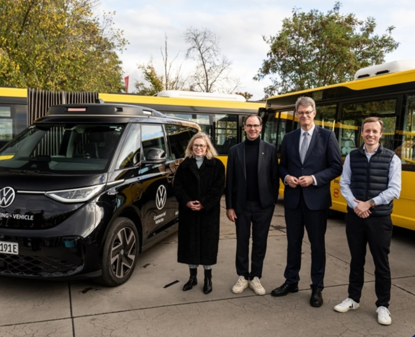 Four people stand in front of a VW autonomous vehicle and yellow buses during a presentation.