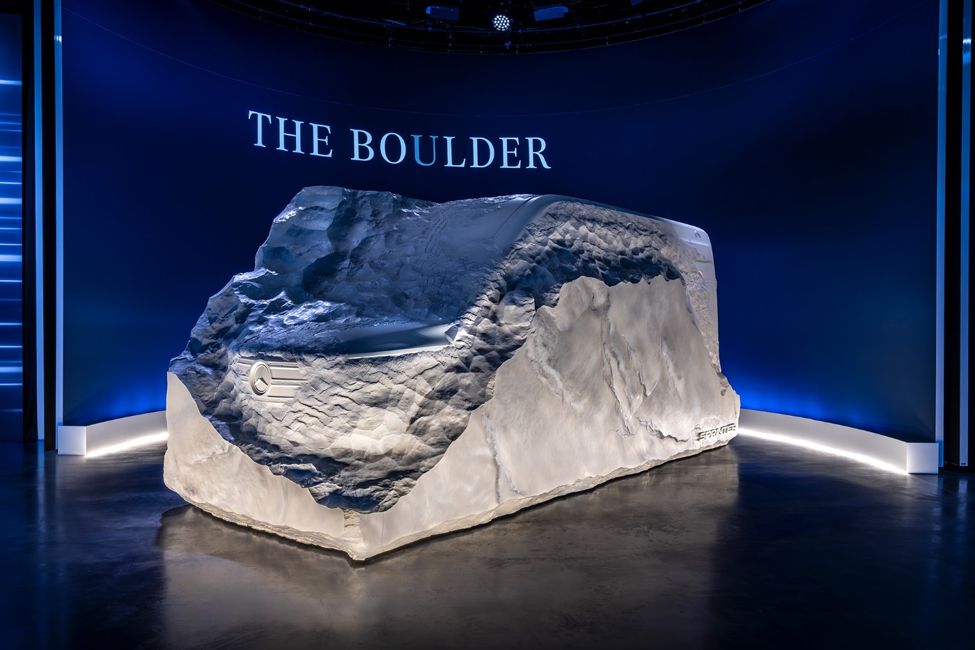 Eine fahrzeugförmige Felsskulptur mit dem Titel „The Boulder“ steht beleuchtet in einem Ausstellungsraum.