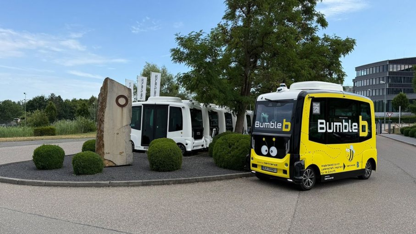Ein gelber autonomer Shuttle mit der Aufschrift „Bumble b“ steht neben weißen Shuttles im Rondell.