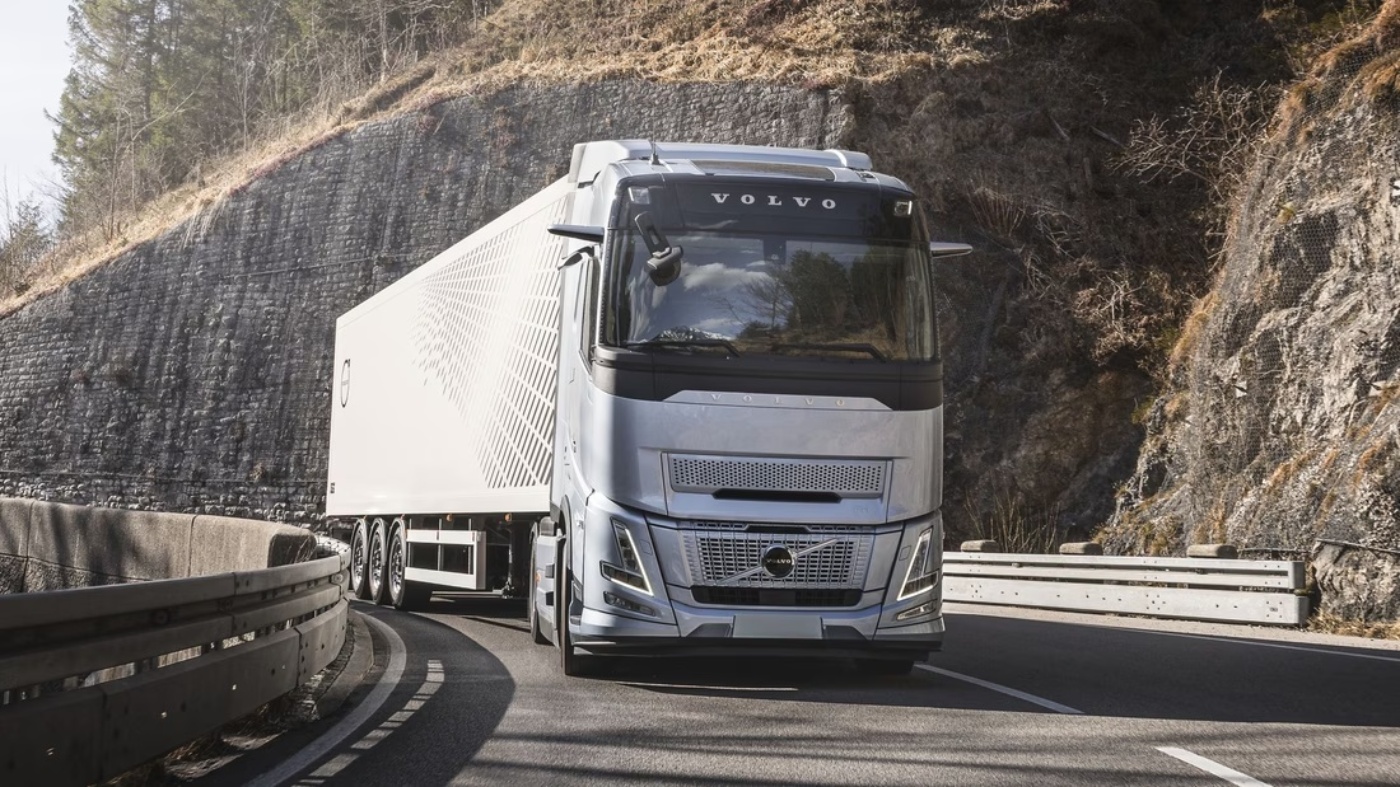 The image shows a silver Volvo semi-truck with a white trailer on a winding road. The truck is the main subject, and it's facing the camera head-on. The road it's traveling on curves slightly, and there are stone walls on either side, suggesting a mountain or rocky terrain.