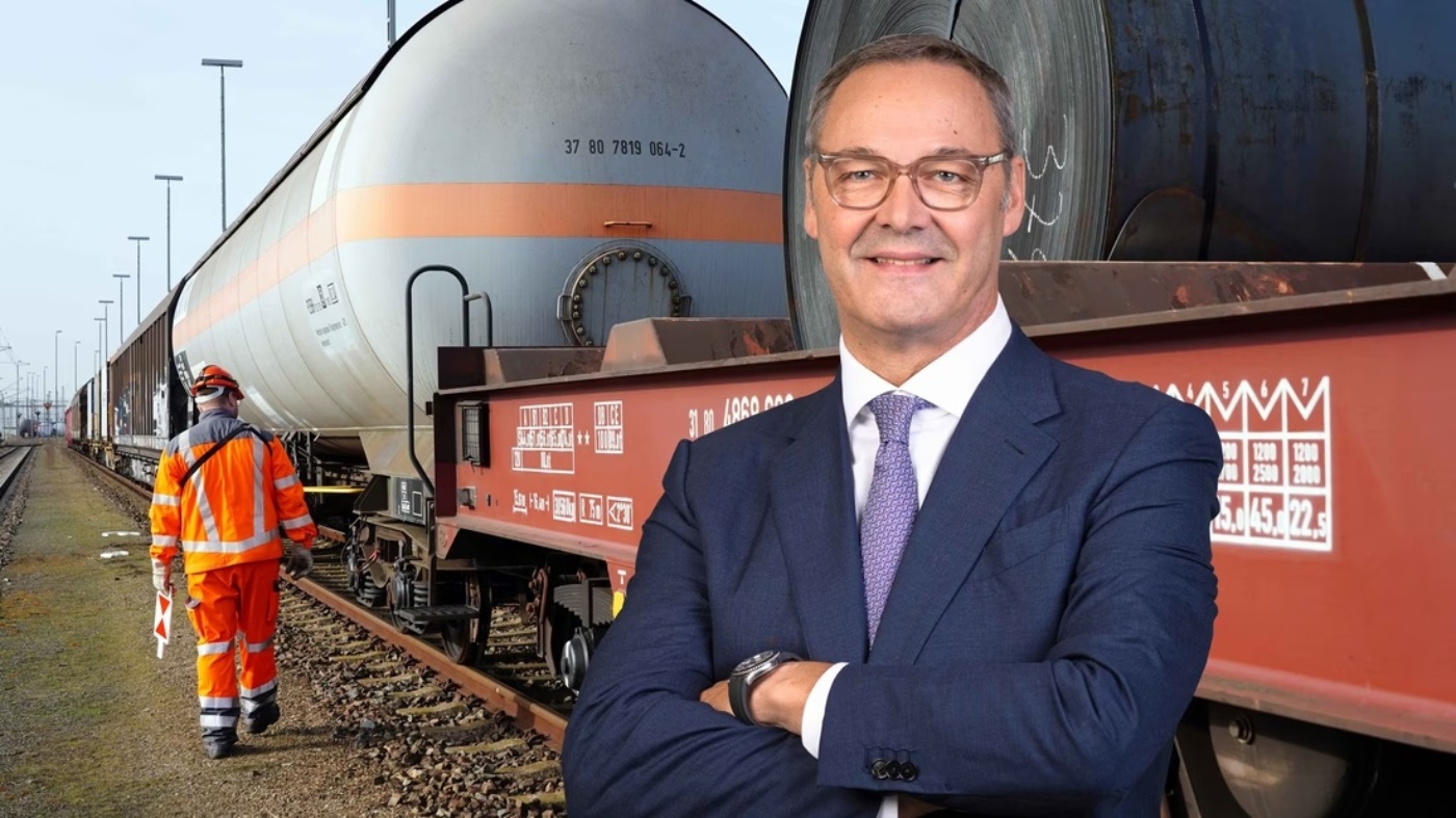 Bernhard Osburg in suit with crossed arms stands in front of freight train with tank cars and a worker in orange safety gear on the railway track.
