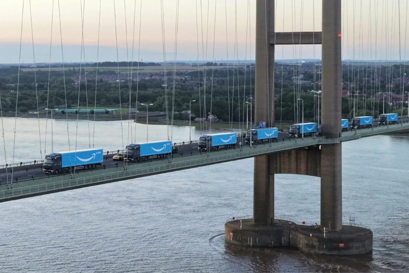 Multiple blue Amazon trucks with white arrows drive on a suspension bridge over a river.