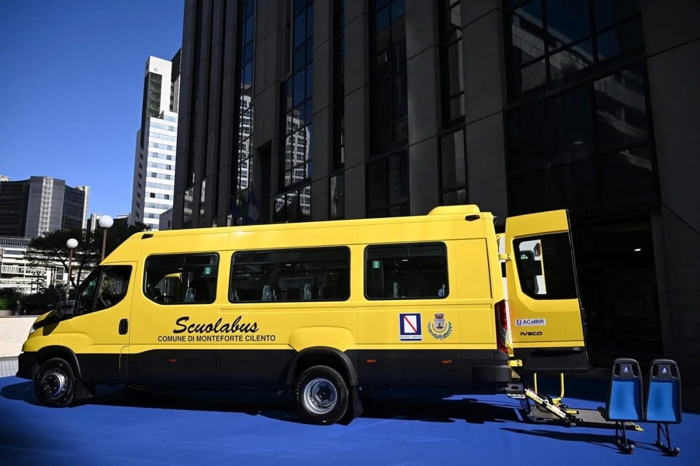 Yellow minibus with open door and extended wheelchair ramp, parked in front of a modern glass building.