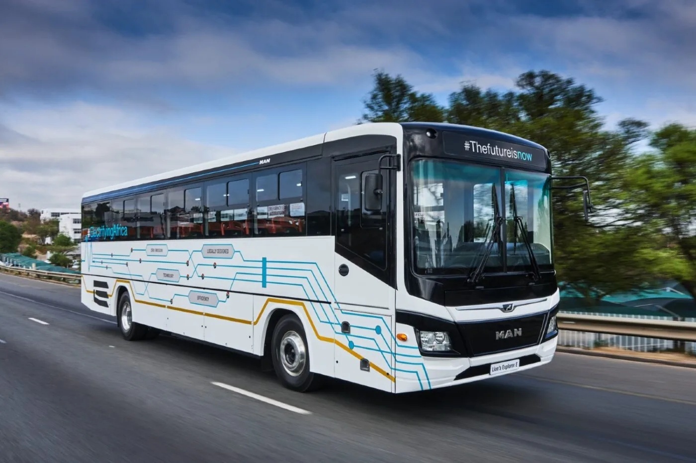 White MAN bus with blue line patterns driving on a road, trees and buildings in the background.