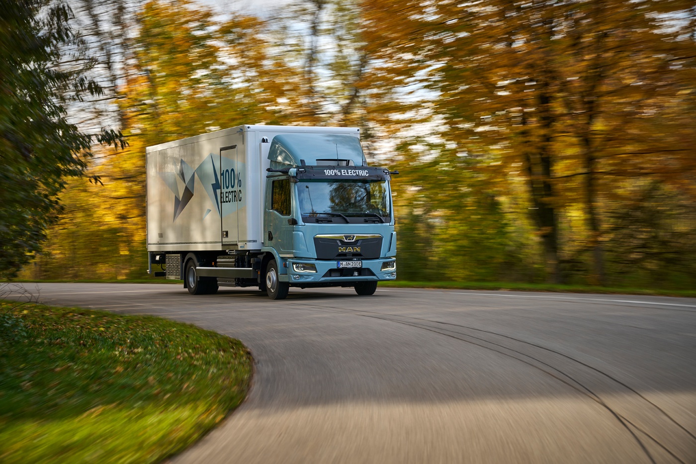 Electric MAN truck with "100% Electric" branding drives on a forest road in autumn.