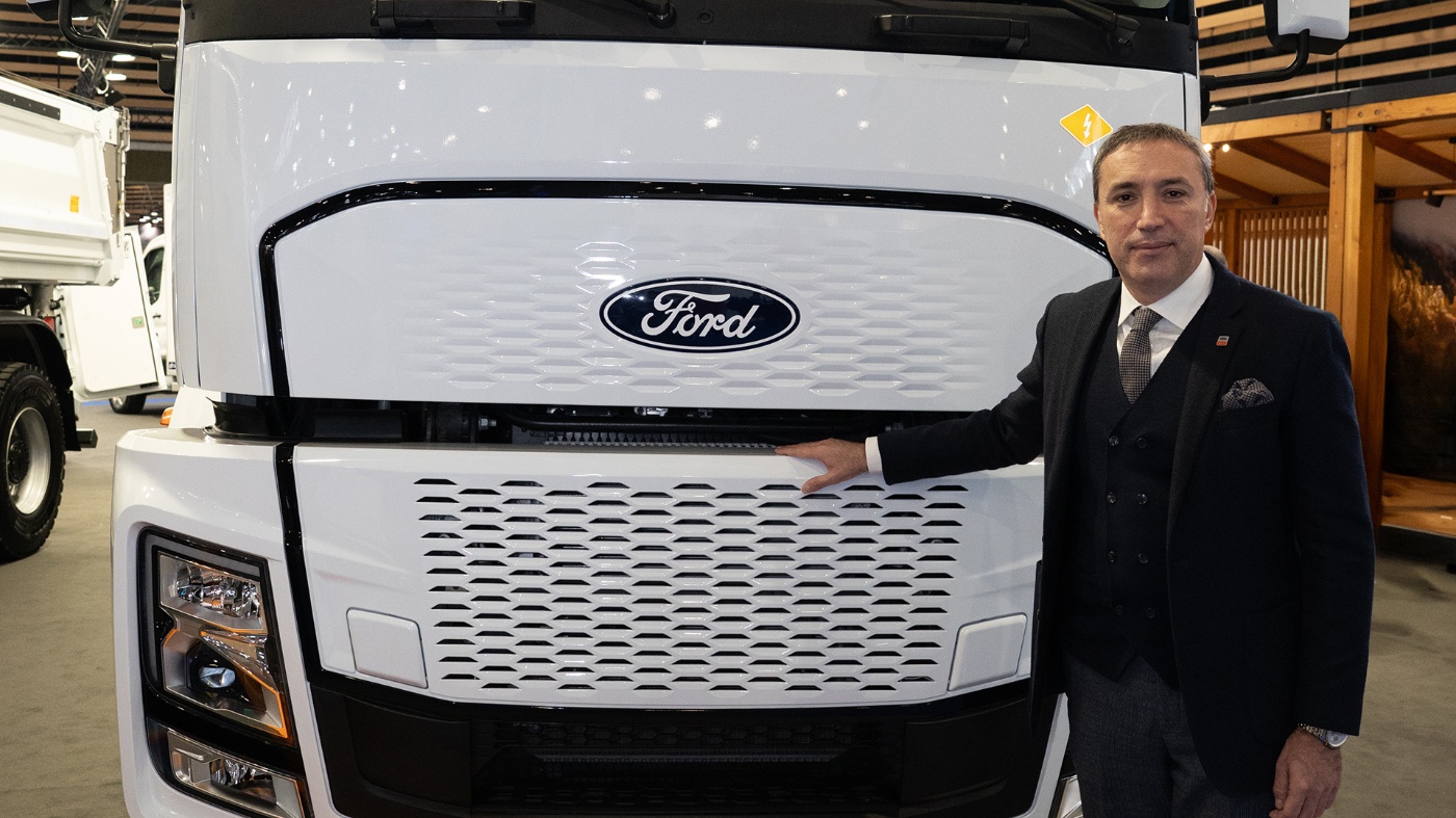 Ein Mann im Anzug steht neben der Front eines großen weißen Ford-Lkw auf einer Messe.