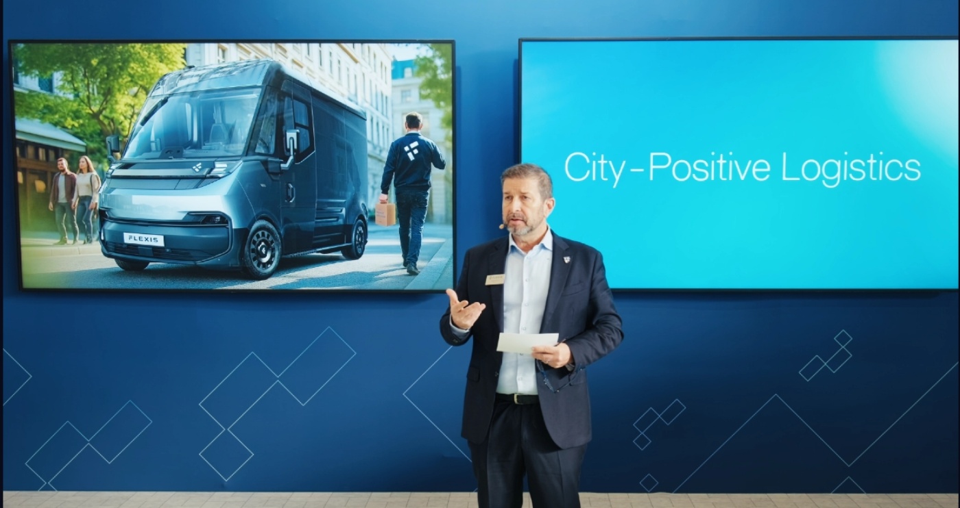 Ein Mann spricht auf einer Bühne vor zwei Bildschirmen mit E-Van und dem Text „City-Positive Logistics“.