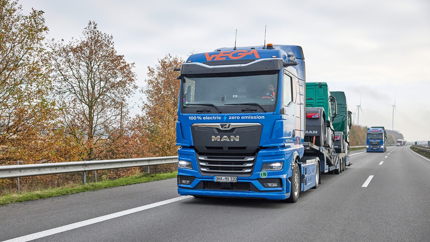 Electric truck transports other trucks on a highway, with wind turbines in the background.