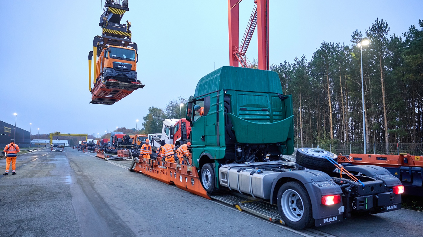 Truck is lifted by crane while workers load vehicles onto a train platform at a logistics terminal.