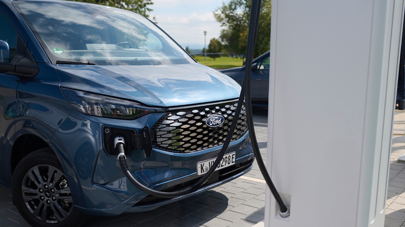 Electric Ford van is plugged into a charging station at a parking area in daylight.