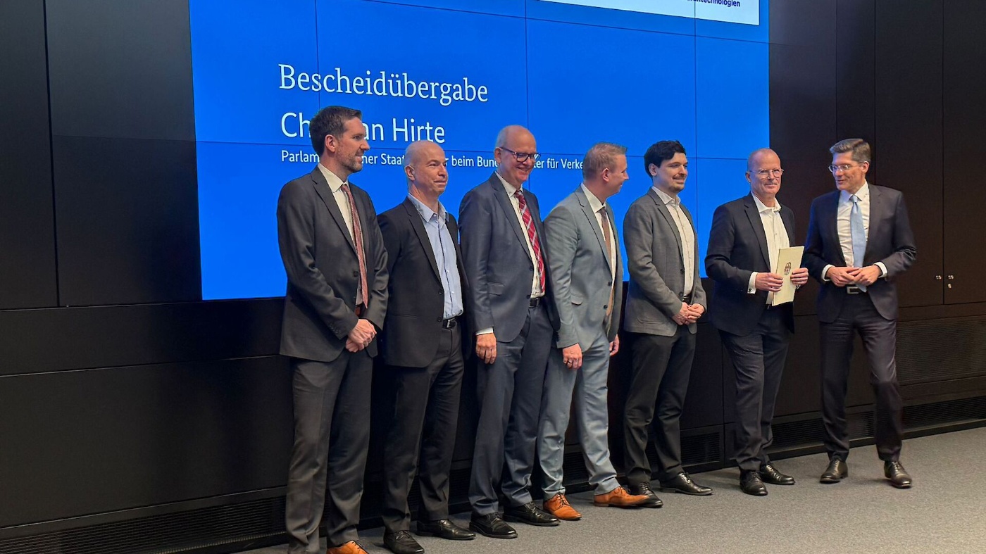 Seven men in suits stand together during a formal handover ceremony in front of a blue screen.