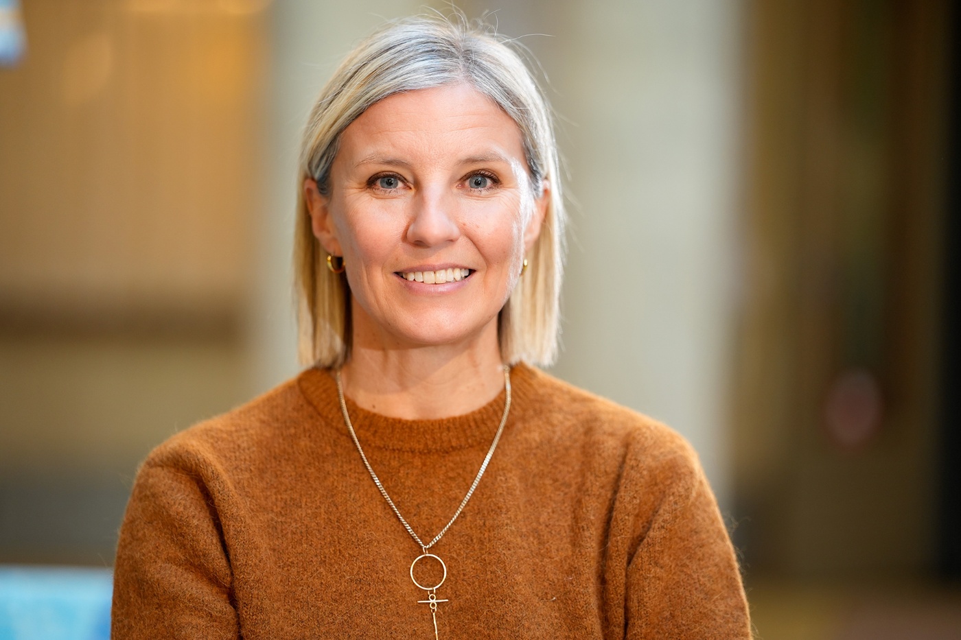 Eine lächelnde Frau mit blondem Haar trägt einen braunen Pullover und eine Kette mit Frauensymbol.