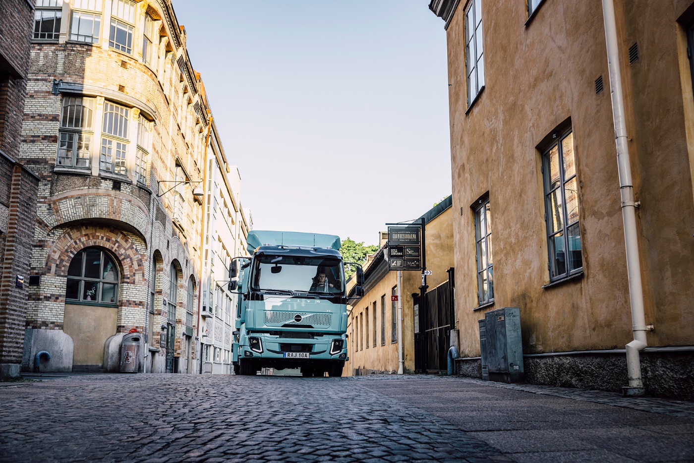 Ein hellblauer Volvo-Lkw fährt durch eine enge Straße mit alten Gebäuden.
