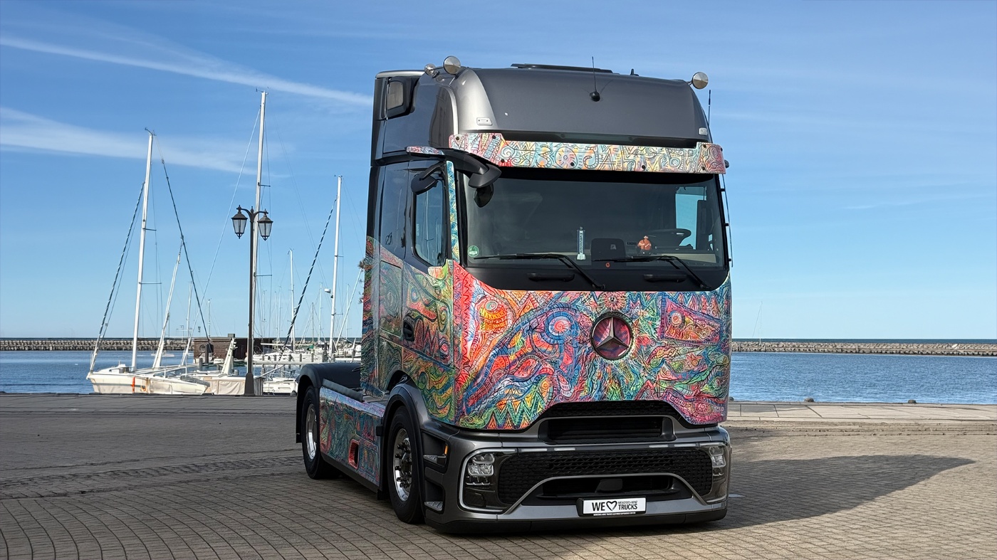 Bunt gestalteter Mercedes-Lkw steht an einem Yachthafen mit Segelbooten im Hintergrund.