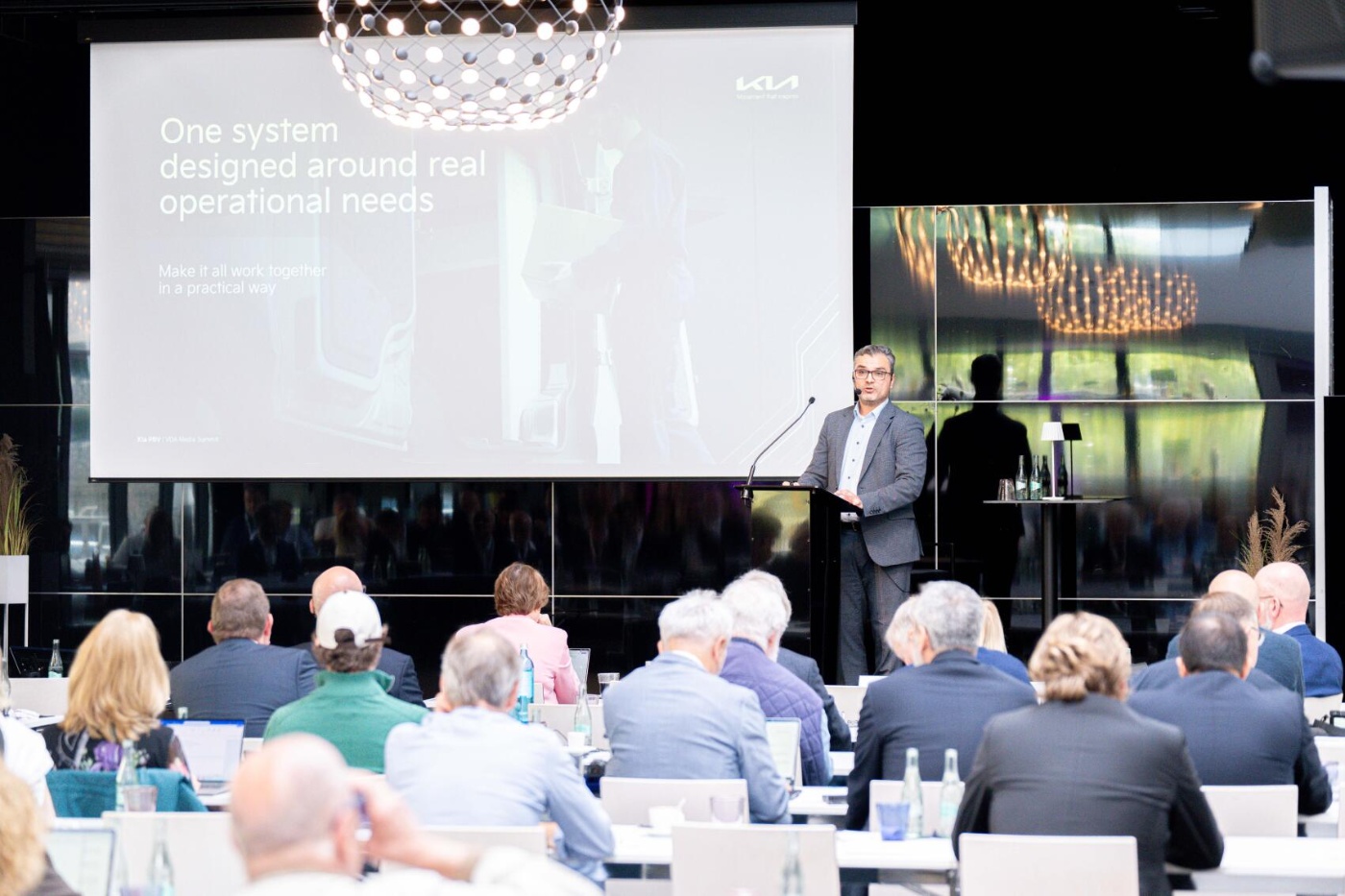 A man speaks at a podium to an audience with a Kia presentation slide behind him.
