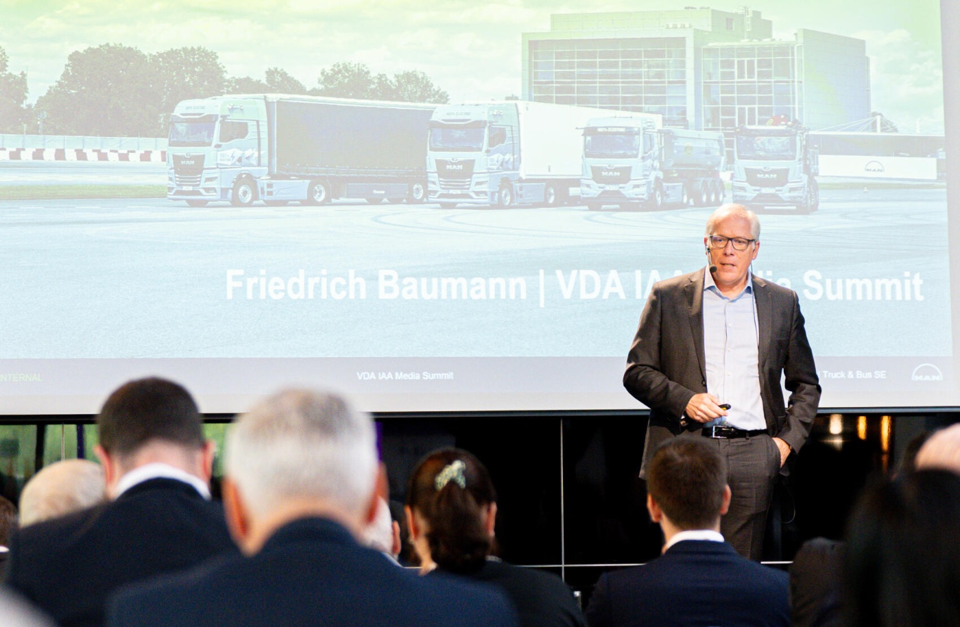 Friedrich Baumann speaks on stage at the VDA IAA Media Summit with trucks on screen.
