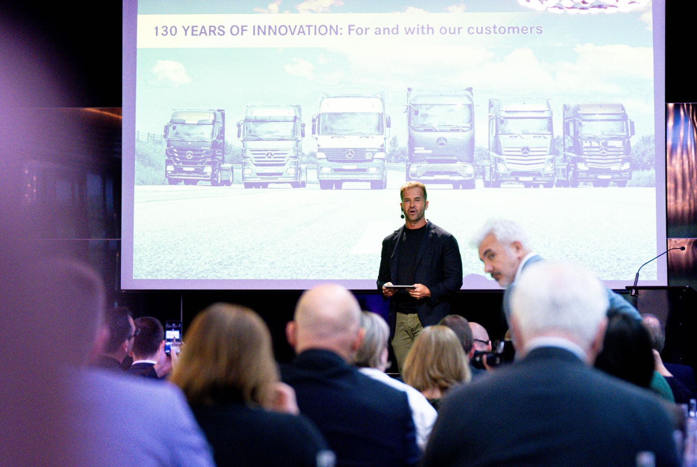 A man presents 130 years of truck innovation on stage before an audience.