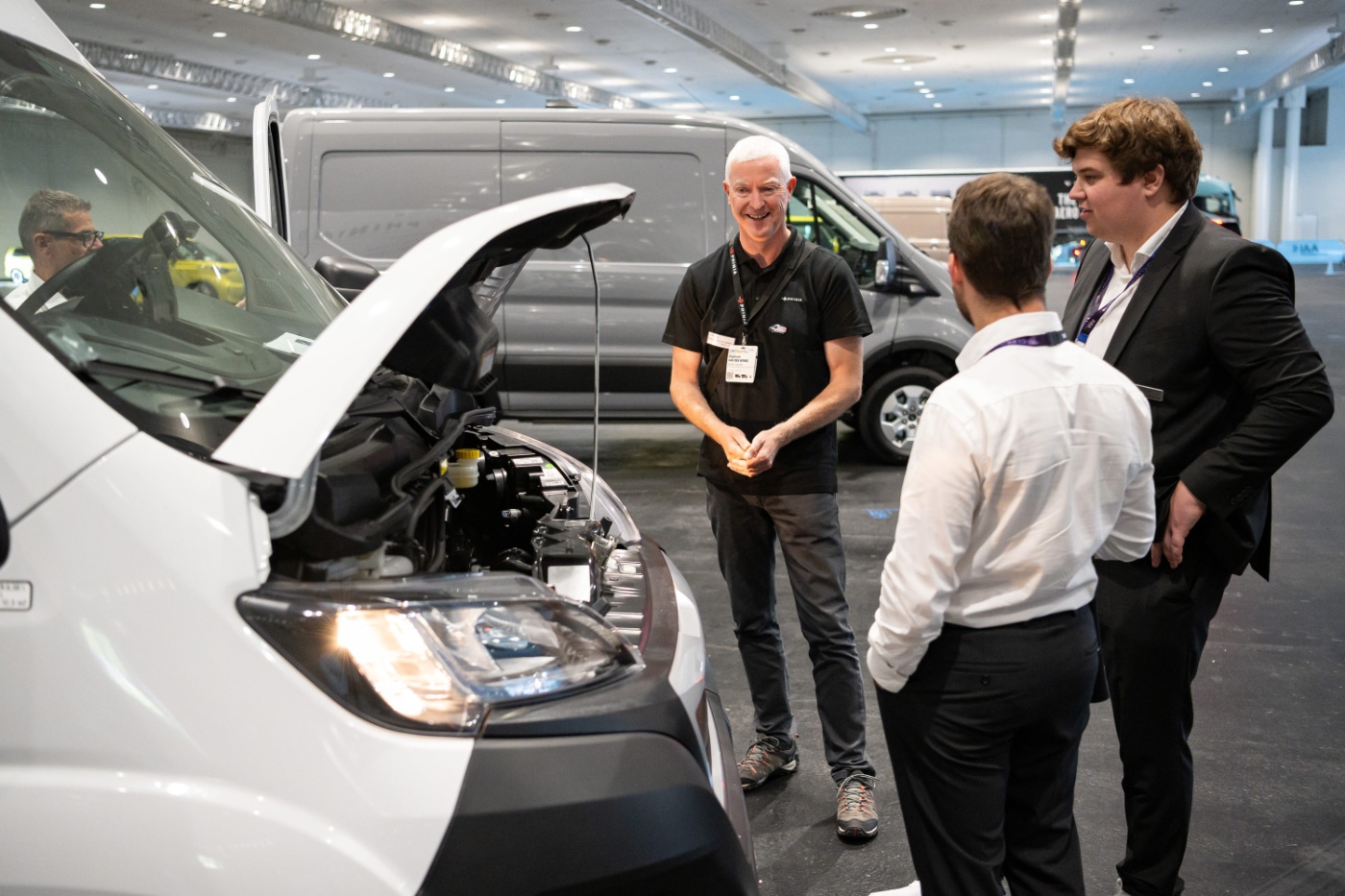 Drei Männer besprechen ein Fahrzeug mit offener Motorhaube auf einer Messe.